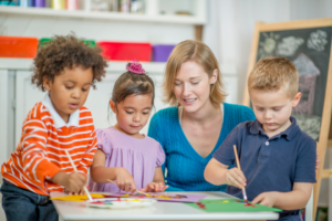 Three children being taught how to paint by a primary school teacher.